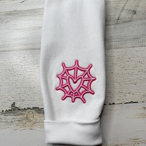 May include: A white long-sleeve garment featuring a pink embroidered design on the sleeve. The embroidery depicts a spider web pattern with a heart shape at the center. The fabric has a smooth texture and a cuffed hem at the bottom of the sleeve.