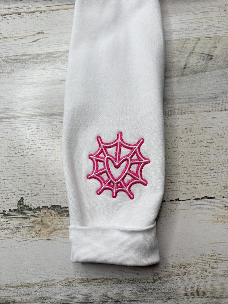 May include: A white long-sleeve garment featuring a pink embroidered design on the sleeve. The embroidery depicts a spider web pattern with a heart shape at the center. The fabric has a smooth texture and a cuffed hem at the bottom of the sleeve.