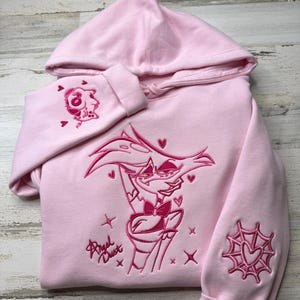 May include: A light pink hooded sweatshirt featuring dark pink embroidery of the character Angel Dust from Hazbin Hotel on the chest, with the name Angel Dust written in script, a spider web heart on one sleeve, and a small character icon on the other.