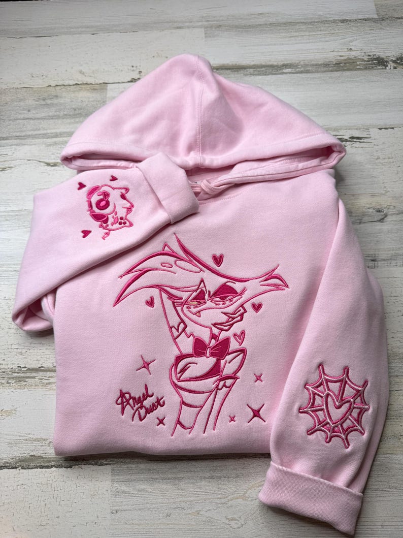 May include: A light pink hooded sweatshirt featuring dark pink embroidery of the character Angel Dust from Hazbin Hotel on the chest, with the name Angel Dust written in script, a spider web heart on one sleeve, and a small character icon on the other.