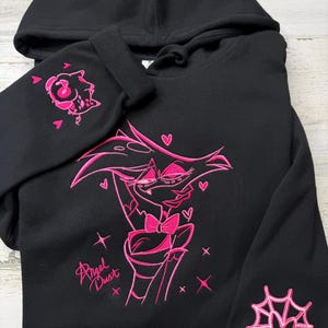 May include: A black hooded sweatshirt featuring bright pink embroidery of the character Angel Dust on the front. The design includes the text Angel Dust, small hearts, and stars. One sleeve features a small character head, and the other a spider web.