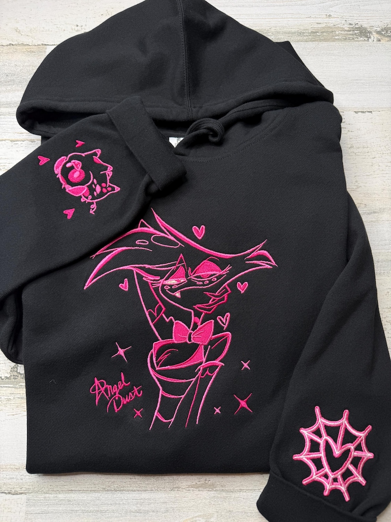 May include: A black hooded sweatshirt featuring bright pink embroidery of the character Angel Dust on the front. The design includes the text Angel Dust, small hearts, and stars. One sleeve features a small character head, and the other a spider web.