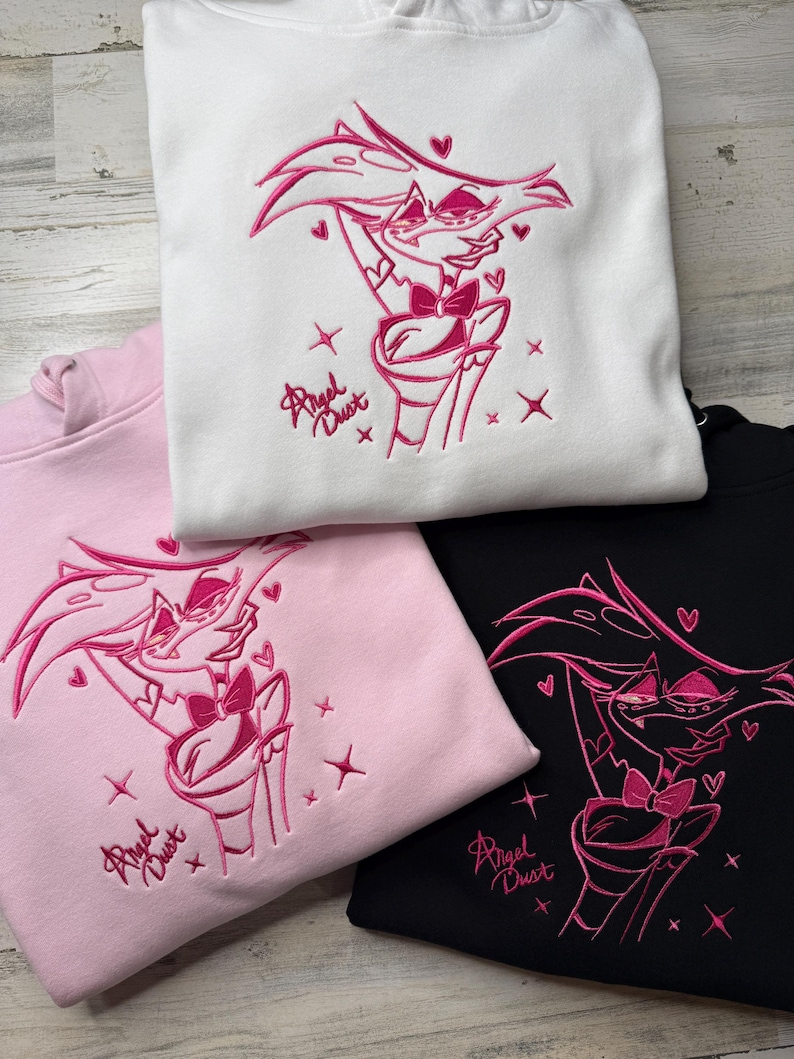 May include: Three hooded sweatshirts in white, light pink, and black, each featuring an embroidered pink graphic of a character with multiple arms, a bow tie, and the text Angel Dust, surrounded by small embroidered hearts and star shapes.