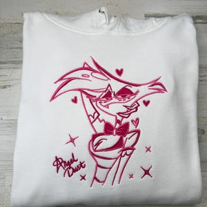 May include: A white hooded sweatshirt featuring a pink embroidered graphic of the character Angel Dust. The design includes the character with a bow tie, surrounded by small hearts and star shapes, with the text Angel Dust written in script.