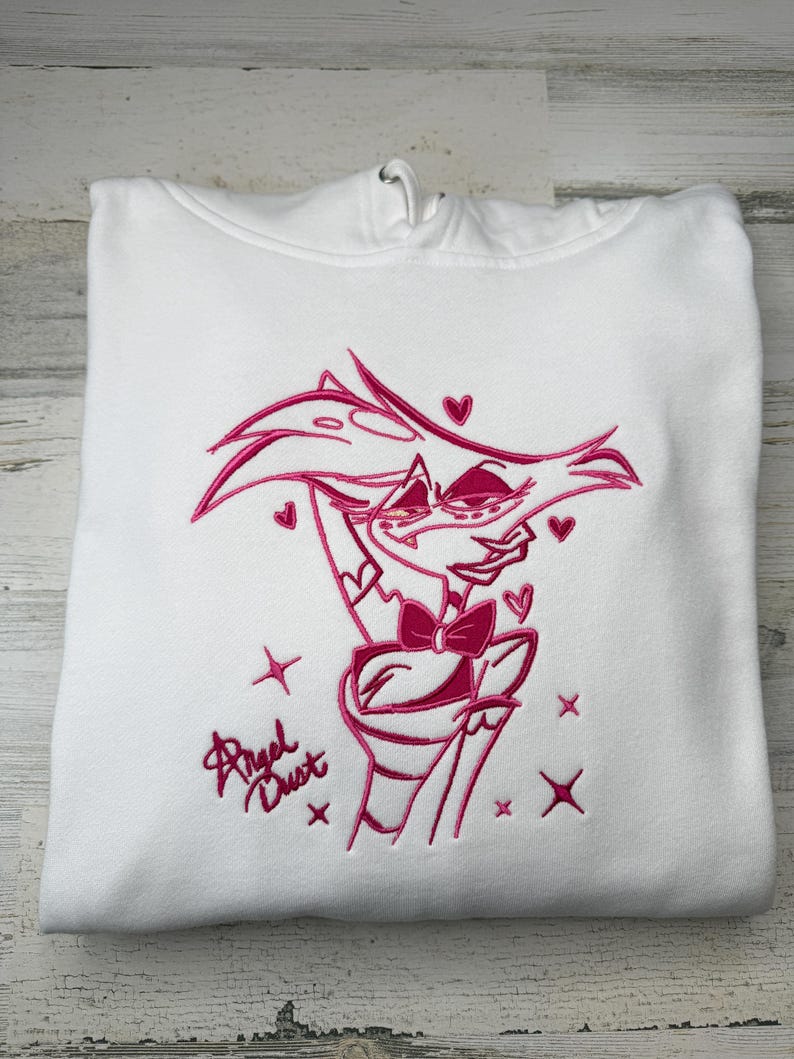 May include: A white hooded sweatshirt featuring a pink embroidered graphic of the character Angel Dust. The design includes the character with a bow tie, surrounded by small hearts and star shapes, with the text Angel Dust written in script.