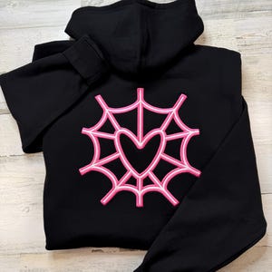 K&ouml;nnte beinhalten: Schwarzer Kapuzenpullover mit einem rosa und wei&szlig;en Spinnennetz-Design mit einem Herz in der Mitte. Der Kapuzenpullover hat eine Kapuze und lange &Auml;rmel. Das Design ist auf der R&uuml;ckseite gestickt.