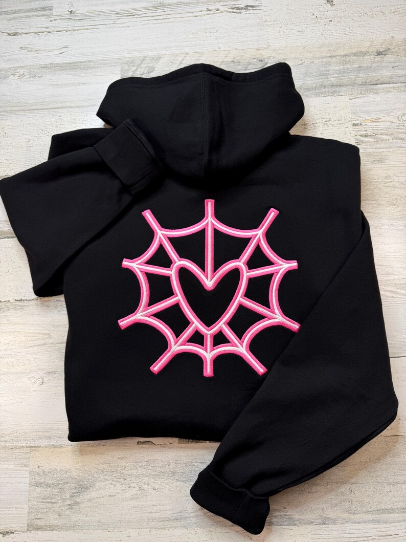 K&ouml;nnte beinhalten: Schwarzer Kapuzenpullover mit einem rosa und wei&szlig;en Spinnennetz-Design mit einem Herz in der Mitte. Der Kapuzenpullover hat eine Kapuze und lange &Auml;rmel. Das Design ist auf der R&uuml;ckseite gestickt.