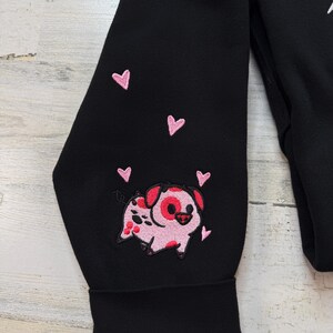 K&ouml;nnte beinhalten: Schwarzer Sweatshirt-&Auml;rmel mit einem gestickten rosa Schwein und rosa Herzen. Das Schwein hat rote Flecken und ein rotes herzf&ouml;rmiges Auge. Die Herzen sind um das Schwein herum verstreut.