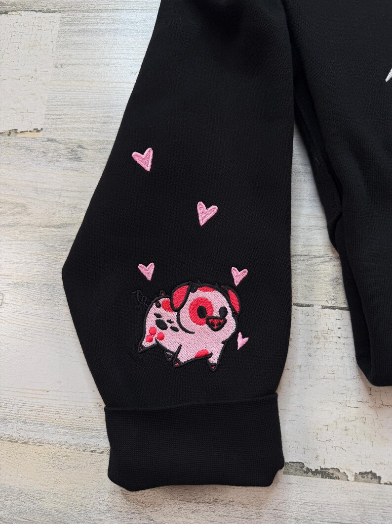 K&ouml;nnte beinhalten: Schwarzer Sweatshirt-&Auml;rmel mit einem gestickten rosa Schwein und rosa Herzen. Das Schwein hat rote Flecken und ein rotes herzf&ouml;rmiges Auge. Die Herzen sind um das Schwein herum verstreut.