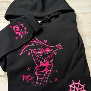 May include: A black hooded sweatshirt featuring pink embroidery of the character Angel Dust from Hazbin Hotel on the front, with the text Angel Dust. The right sleeve displays a small embroidered character head, and the left sleeve has a spider web.