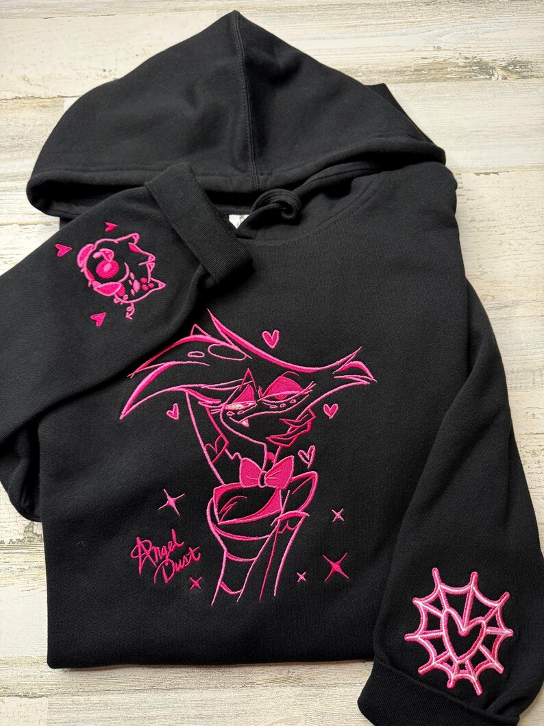 May include: A black hooded sweatshirt featuring pink embroidery of the character Angel Dust from Hazbin Hotel on the front, with the text Angel Dust. The right sleeve displays a small embroidered character head, and the left sleeve has a spider web.