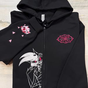 K&ouml;nnte beinhalten: Schwarzer Kapuzenpullover mit Rei&szlig;verschluss und Kapuze. Der linke &Auml;rmel zeigt ein rosa und rotes Stickdesign mit rosa Herzen. Auf der Vorderseite befindet sich eine wei&szlig;-rosa Grafik einer Figur und der Text "HAZBIN HOTEL" in Rosa.