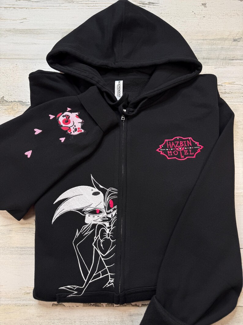 K&ouml;nnte beinhalten: Schwarzer Kapuzenpullover mit Rei&szlig;verschluss und Kapuze. Der linke &Auml;rmel zeigt ein rosa und rotes Stickdesign mit rosa Herzen. Auf der Vorderseite befindet sich eine wei&szlig;-rosa Grafik einer Figur und der Text "HAZBIN HOTEL" in Rosa.