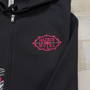 K&ouml;nnte beinhalten: Schwarzer Kapuzenpullover mit Rei&szlig;verschluss und einem rosa gestickten Design. Das Design zeigt die Worte "HAZBIN HOTEL" in einer stilisierten Schriftart mit dekorativen Elementen. Der Hoodie hat einen durchgehenden Rei&szlig;verschluss.