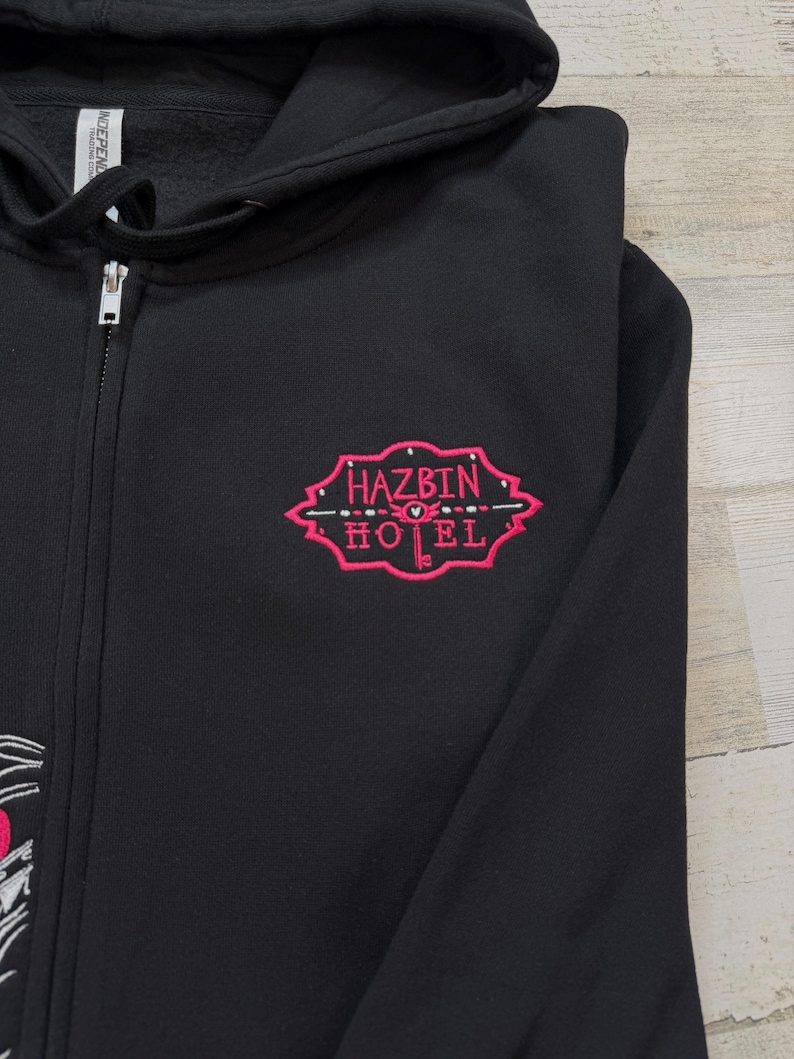 K&ouml;nnte beinhalten: Schwarzer Kapuzenpullover mit Rei&szlig;verschluss und einem rosa gestickten Design. Das Design zeigt die Worte "HAZBIN HOTEL" in einer stilisierten Schriftart mit dekorativen Elementen. Der Hoodie hat einen durchgehenden Rei&szlig;verschluss.
