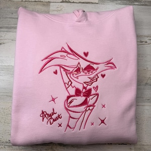 May include: A light pink hooded sweatshirt featuring a dark pink embroidered graphic of the character Angel Dust. The design includes the character's portrait, small heart shapes, four-pointed star accents, and the text Angel Dust in script.