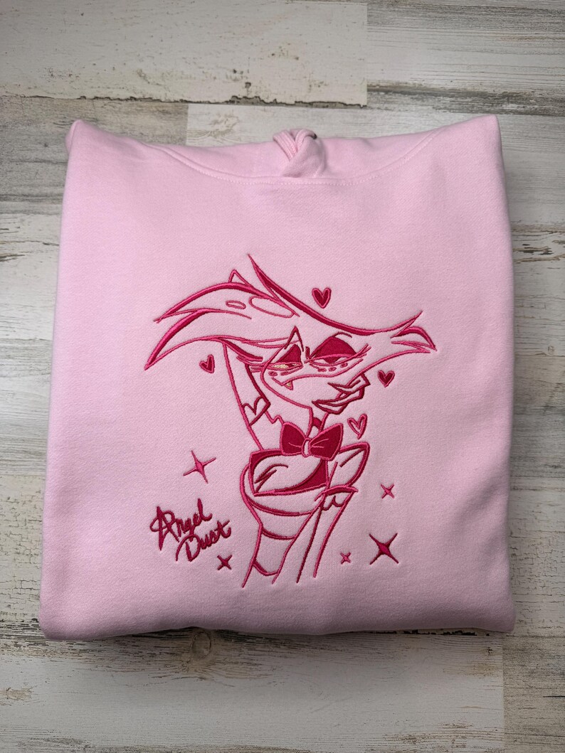 May include: A light pink hooded sweatshirt featuring a dark pink embroidered graphic of the character Angel Dust. The design includes the character's portrait, small heart shapes, four-pointed star accents, and the text Angel Dust in script.