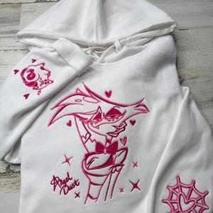 May include: A white hooded sweatshirt featuring pink embroidery of the character Angel Dust on the chest, with the text Angel Dust below. The right sleeve displays a small character icon, and the left sleeve features a pink spider web design.