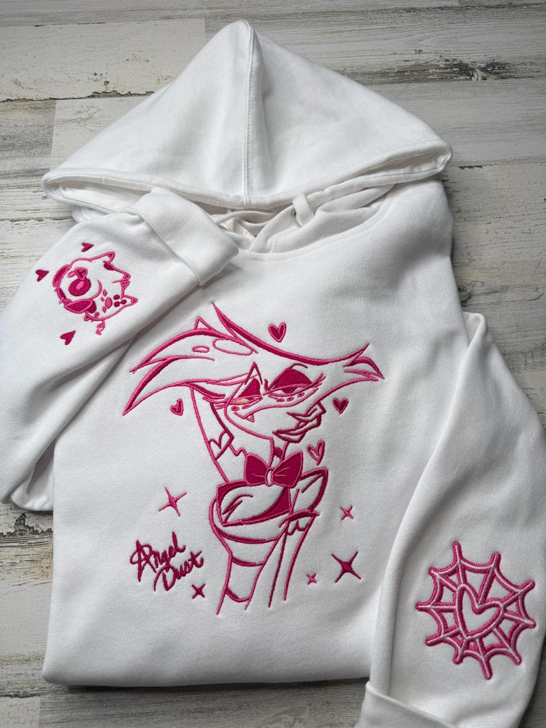 May include: A white hooded sweatshirt featuring pink embroidery of the character Angel Dust on the chest, with the text Angel Dust below. The right sleeve displays a small character icon, and the left sleeve features a pink spider web design.