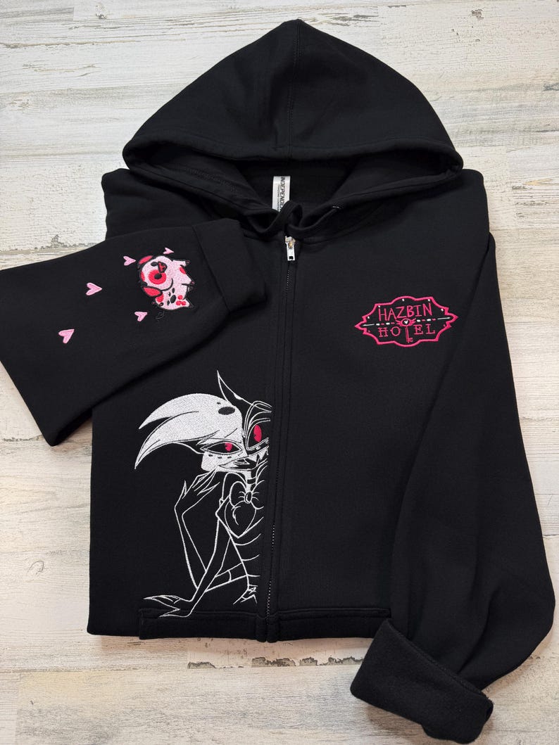 K&ouml;nnte beinhalten: Schwarzer Kapuzenpullover mit Rei&szlig;verschluss, Kapuze und langen &Auml;rmeln. Der linke &Auml;rmel zeigt eine rosa-wei&szlig;e Cartoonfigur und rosa Herzen. Auf der Vorderseite ist eine schwarz-wei&szlig;e Illustration und der Text "HAZBIN HOTEL" in Rosa zu sehen.