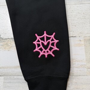 May include: A black long-sleeved garment featuring an embroidered design on the sleeve. The embroidery consists of a bright pink spider web pattern surrounding a central heart shape, with a ribbed cuff at the bottom of the sleeve.
