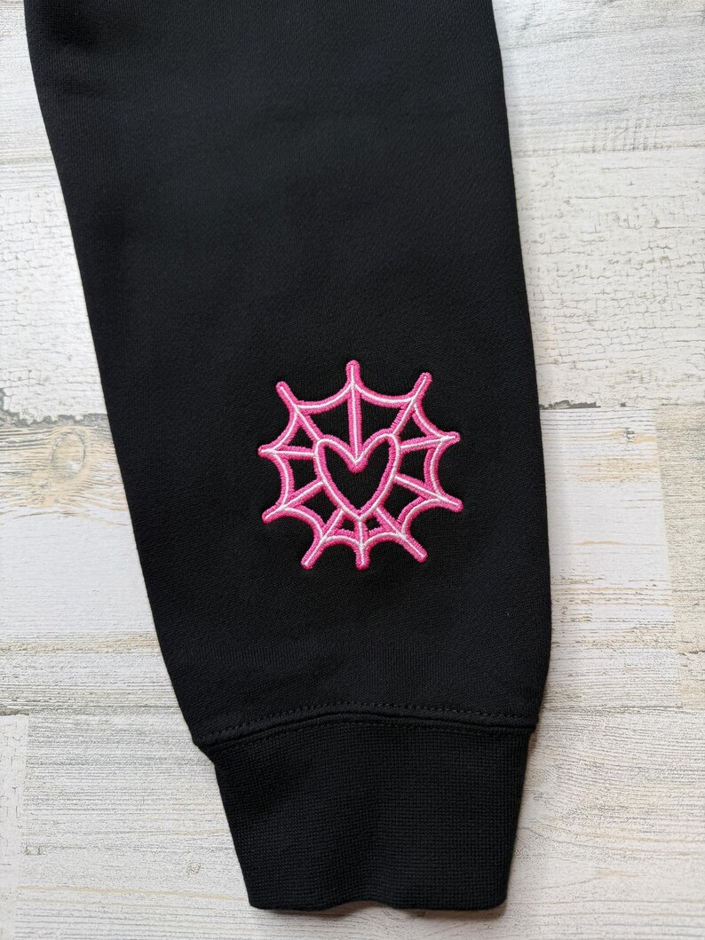 May include: A black long-sleeved garment featuring an embroidered design on the sleeve. The embroidery consists of a bright pink spider web pattern surrounding a central heart shape, with a ribbed cuff at the bottom of the sleeve.
