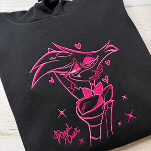 May include: A black hooded sweatshirt featuring a vibrant pink embroidered graphic of the character Angel Dust. The design includes the character with a bow tie, surrounded by small hearts and star accents, with the text Angel Dust below.