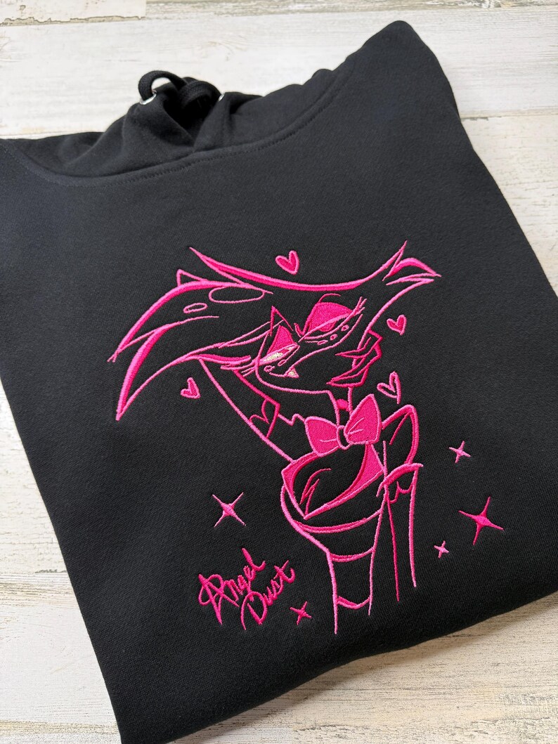 May include: A black hooded sweatshirt featuring a vibrant pink embroidered graphic of the character Angel Dust. The design includes the character with a bow tie, surrounded by small hearts and star accents, with the text Angel Dust below.