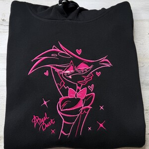 May include: A black hooded sweatshirt featuring a vibrant pink embroidered graphic of the character Angel Dust. The design includes the character with a bow tie, surrounded by small hearts and star shapes, with the text Angel Dust written below.