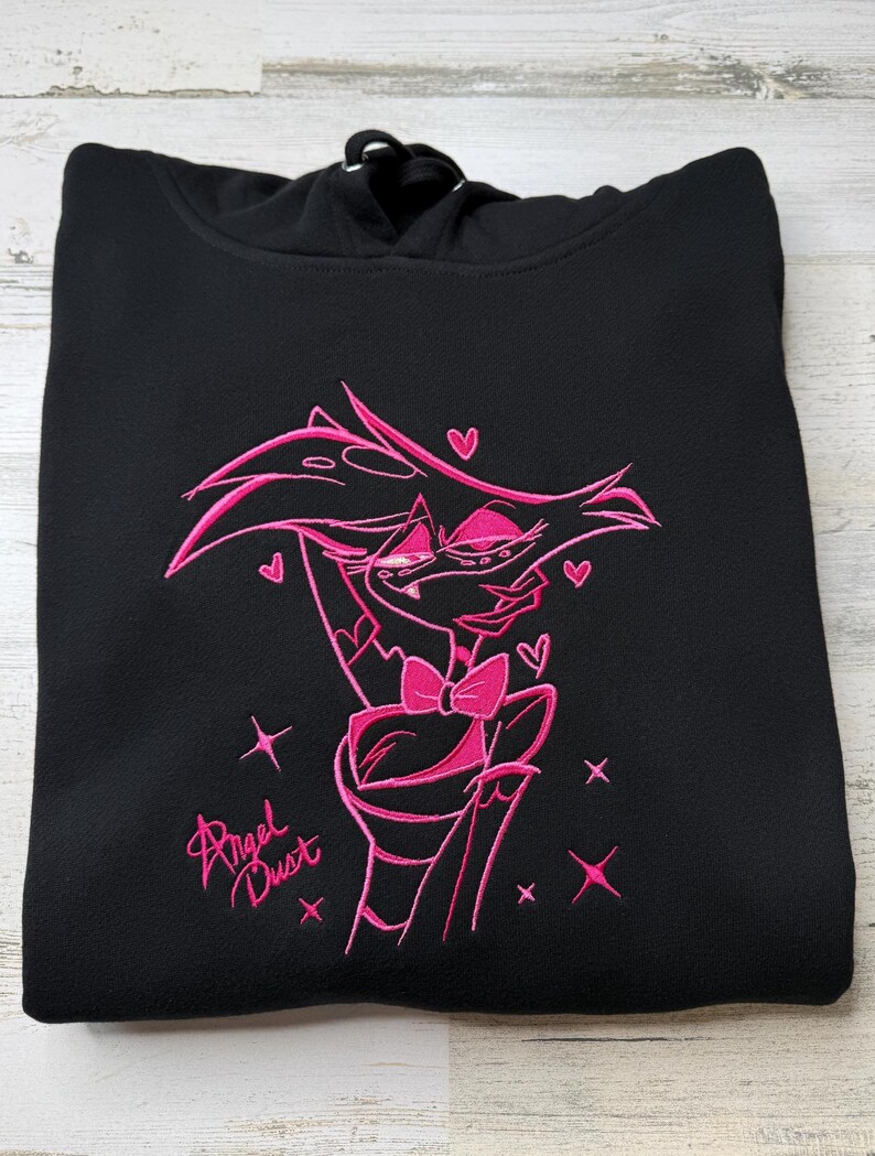May include: A black hooded sweatshirt featuring a vibrant pink embroidered graphic of the character Angel Dust. The design includes the character with a bow tie, surrounded by small hearts and star shapes, with the text Angel Dust written below.