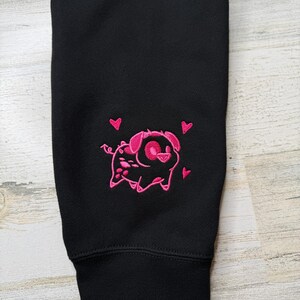 May include: A black long-sleeve garment featuring an embroidered design of a small pig with a curly tail and pointed ears. The pig is outlined in bright pink thread and surrounded by three small pink hearts, positioned above the ribbed cuff.