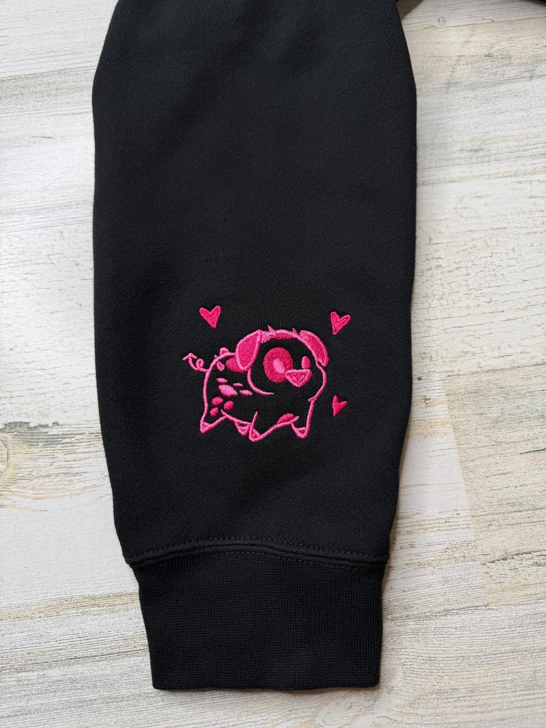 May include: A black long-sleeve garment featuring an embroidered design of a small pig with a curly tail and pointed ears. The pig is outlined in bright pink thread and surrounded by three small pink hearts, positioned above the ribbed cuff.