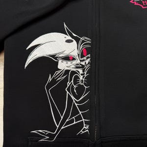 K&ouml;nnte beinhalten: Schwarze Jacke mit Rei&szlig;verschluss und wei&szlig;-roter Stickerei eines Cartoon-Charakters. Die Jacke zeigt eine detaillierte Illustration eines Charakters mit wei&szlig;em Gesicht, roten Augen und einer Fliege. Mit einem rosa Logo.