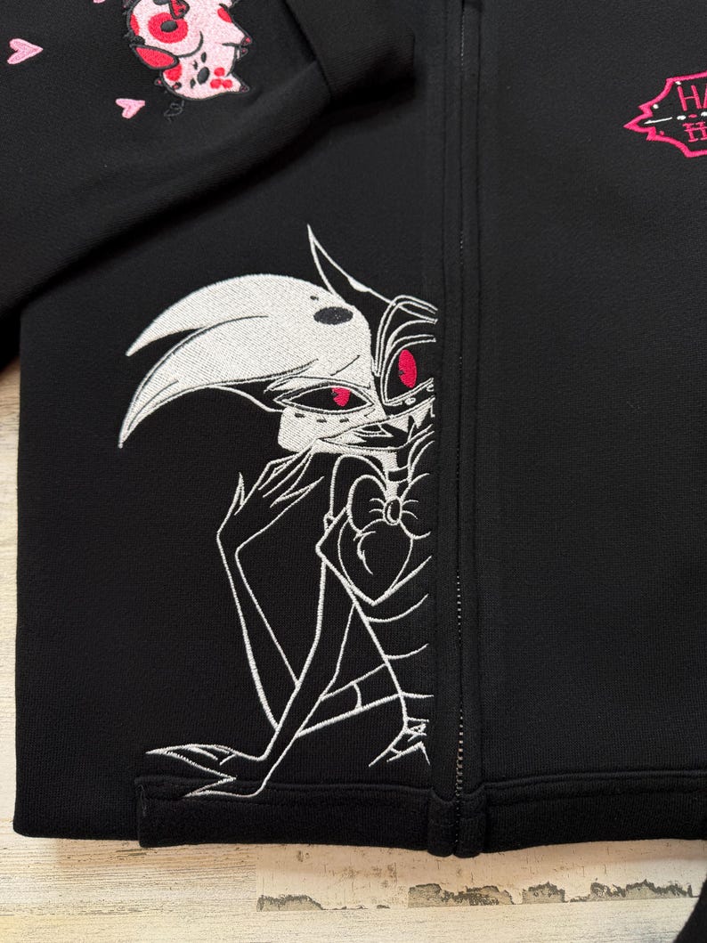 K&ouml;nnte beinhalten: Schwarze Jacke mit Rei&szlig;verschluss und wei&szlig;-roter Stickerei eines Cartoon-Charakters. Die Jacke zeigt eine detaillierte Illustration eines Charakters mit wei&szlig;em Gesicht, roten Augen und einer Fliege. Mit einem rosa Logo.