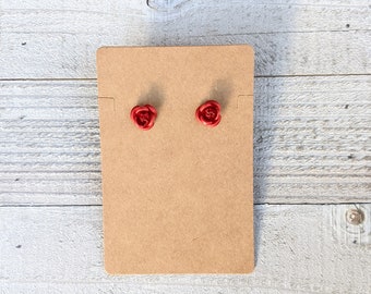 May include: Two small, red rose-shaped stud earrings displayed on a brown cardboard backing. The earrings are a vibrant red color and have detailed petals. The background is a light-colored, weathered wood surface.