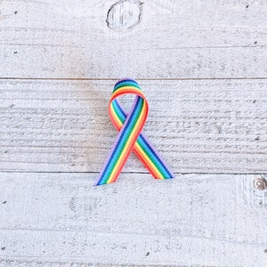 May include: A rainbow ribbon, fashioned into a looped bow, is positioned on a white wooden background. The ribbon showcases a spectrum of colours: red, orange, yellow, green, blue, and purple, symbolising the LGBTQ+ pride flag.
