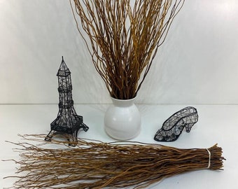 May include: A collection of decorative items, including a bundle of brown twigs, a white vase filled with twigs, and two black wireframe sculptures of the Eiffel Tower and a high-heeled shoe. The items are arranged on a white surface.