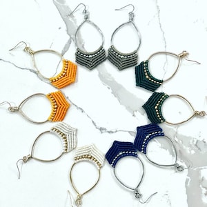 May include: A set of eight pairs of handmade macrame earrings. The earrings are made with a variety of colors, including orange, white, green, blue, and gray. Each pair of earrings features a unique design.