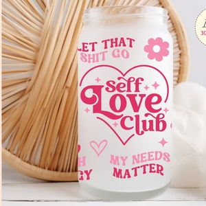 Puede incluir: Un vaso de sublimación blanco con texto rosa que dice "Let that shit go" y "Self Love Club". El vaso también tiene un corazón rosa con una flor rosa en la parte superior. La parte inferior del vaso dice "My Needs Matter".