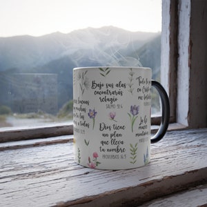 May include: White ceramic mug with a black handle. The mug has a floral design and inspirational quotes in Spanish. The quotes include "Bajo sus alas encontraras refugio" (Psalm 91:4), "Dios tiene un plan que lleva tu nombre" (Proverbs 16:9), "Todo lo puedo en Cristo que me fortalece" (Philippians 4:13), and "La lo mueve monta&ntilde;as" (Matthew 17:20).