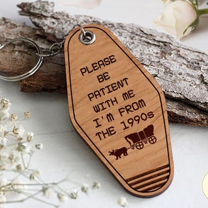 May include: Wooden keychain shaped like a hotel key with the text "Please be patient with me I'm from the 1990s" and a small image of a covered wagon.