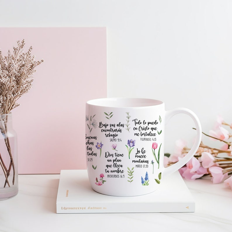 May include: White ceramic mug with floral designs and inspirational quotes in Spanish. The quotes include "Bajo sus alas encontraras refugio" (Psalm 9:4), "Todo lo puedo en Cristo que me fortalece" (Philippians 4:13), "Dios tiene un plan que lleva tu nombre" (Proverbs 16:9), and "Yo le muevo monta&ntilde;as" (Matthew 17:20).