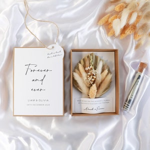 Puede incluir: Un juego de regalo de boda con una tarjeta con el texto "Forever and ever" y nombres, una caja con flores secas y un tubo de vidrio con un corcho. El juego está sobre una superficie de satén blanco.
