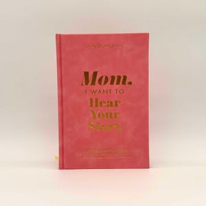 May include: A coral-colored journal with gold lettering that reads "Mom, I want to hear your story." The journal has a soft, textured cover and a gold spine. The title "Family Keepsake Journal" is at the top.