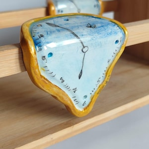 May include: A surrealist-style clock sculpture with a melting face, painted in light blue with black hands and gold trim. The clock is mounted on a wooden shelf, evoking the style of Salvador Dalí.