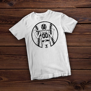 Peut inclure: T-shirt blanc avec un graphique noir et blanc d'un joueur de baseball tenant une batte. Le graphique comprend un ballon de baseball avec des coutures et le texte "YOUR NAME" et "00".