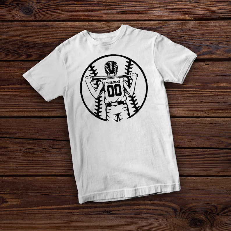 Peut inclure: T-shirt blanc avec un graphique noir et blanc d'un joueur de baseball tenant une batte. Le graphique comprend un ballon de baseball avec des coutures et le texte "YOUR NAME" et "00".