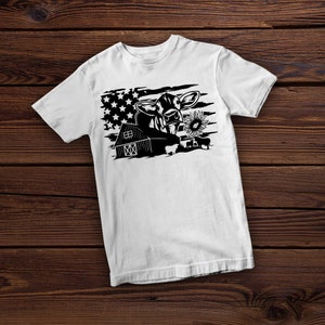 May include: White t-shirt with a black and white graphic of a cow with a sunflower in its mouth, a barn, and an American flag in the background.