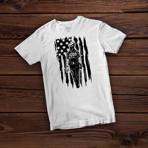 Op de afbeelding: Wit T-shirt met een zwart-witte afbeelding van een politieagent die voor een verscheurde Amerikaanse vlag staat. De agent draagt een hoed en heeft het woord "POLICE" op zijn borst.