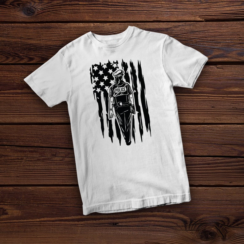 Op de afbeelding: Wit T-shirt met een zwart-witte afbeelding van een politieagent die voor een verscheurde Amerikaanse vlag staat. De agent draagt een hoed en heeft het woord "POLICE" op zijn borst.
